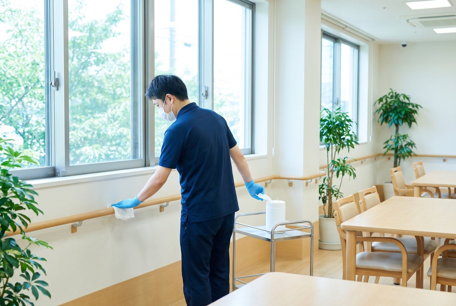 介護施設の明るい共用スペースで、マスクと手袋を着けたスタッフが、消毒用のシートで手すりを拭く様子